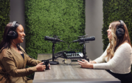 Two women sit at a table engaged in a podcast recording, surrounded by lush green wall decor and equipped with microphones and audio equipment.